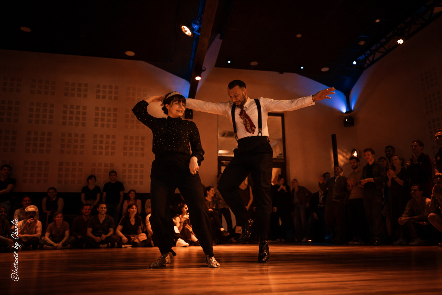 Couple de Lindy Hop en swing out, bras tendus, devant les danseurs assis
